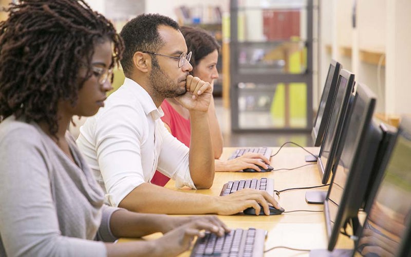 Multiracial group of students training in computer class. Line of man and women in casual sitting at table, using desktops, typing, looking at monitor. Training center concept